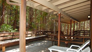 A covered wooden deck with benches and lounge chairs, surrounded by trees.