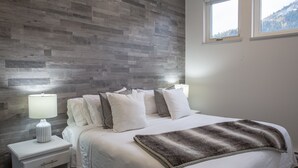 Ein Schlafzimmer mit Holzwand, einem Bett mit weißer Bettwäsche, einem Nachttisch mit Lampe und einem Fenster mit Blick auf die Berge.