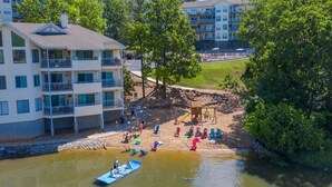 Vista a la orilla del río de una zona residencial con un edificio de varios pisos, un parque infantil y personas disfrutando de actividades acuáticas.