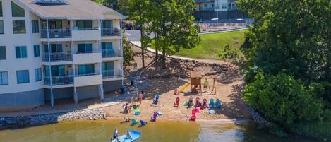 A riverside view of a residential area with a multi-story building, a playground, and people engaging in water activities.