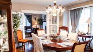 Une salle à manger avec une table en bois, des chaises et un lustre. On aperçoit une plante dans un coin et une cheminée au fond.