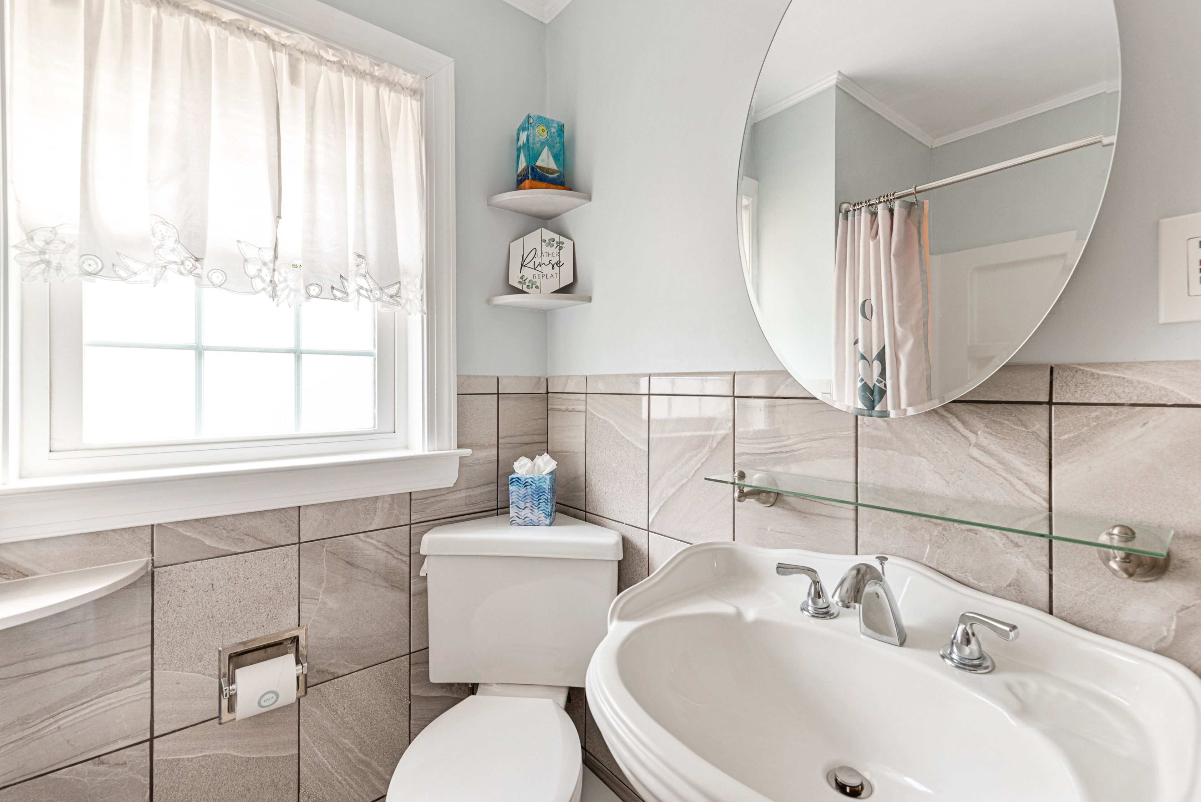 A bathroom with a toilet, sink, and bathtub. There is a window with curtains, a shelf with a decorative item, and a mirror above the bathtub.