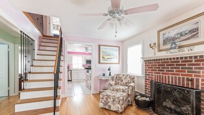 A staircase with a wooden finish and black railings, leading to a kitchen area with white cabinets and a pink wall. A floral armchair is placed near a fireplace with a brick surround.
