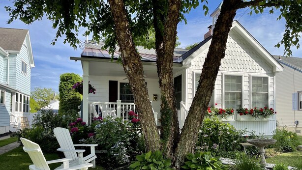 A house with a porch, two trees, and a garden with plants and flowers.