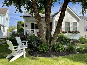 A house with a porch, two trees, and a garden with plants and flowers.