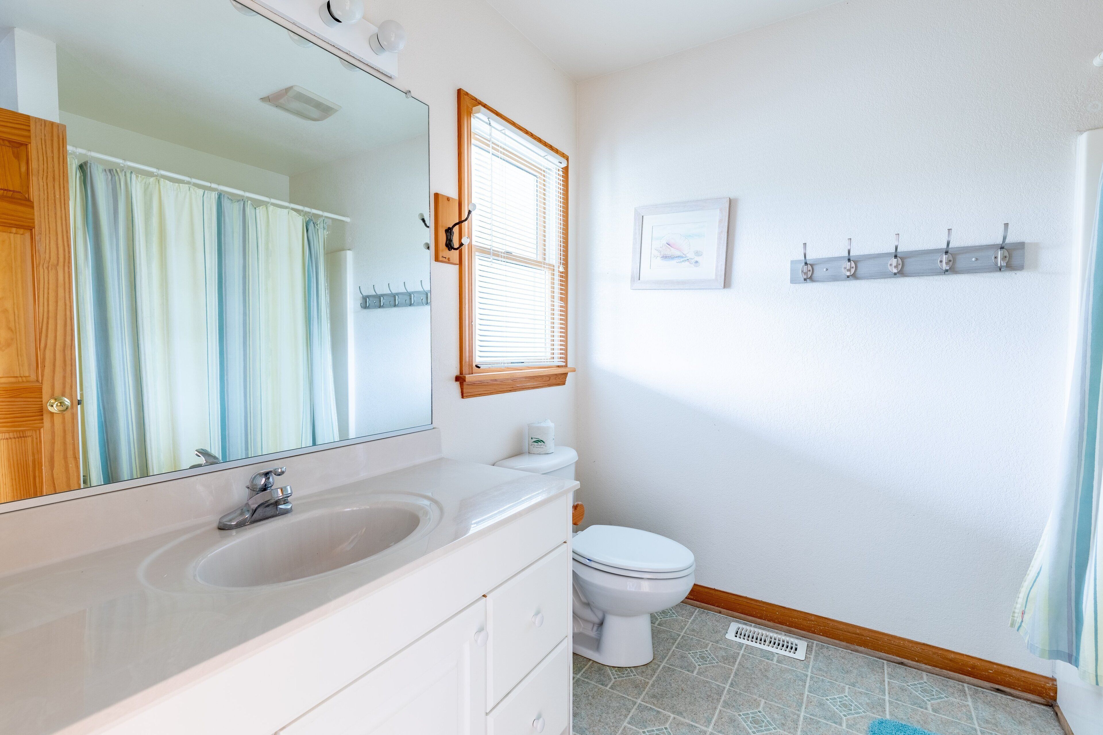A bathroom with a white sink, mirror, and toilet. There is a window with blinds, a towel rack, and a picture on the wall.