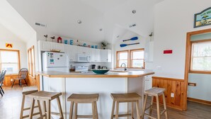 Eine Küche mit weißen Schränken, einer zentralen Kochinsel mit Sitzgelegenheiten, Holzböden und Blick ins Freie durch ein Fenster.