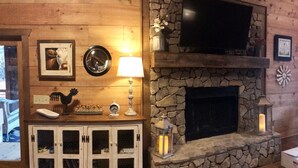 A cozy living room with a stone fireplace, a mounted television, and a wooden cabinet with glass doors. - Luxury cabin! Designer decorated.  Incredible views! Amazing sunrise! Secluded!  (Blue Ridge)