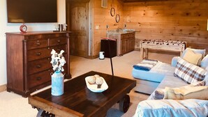A cozy living room with a wooden floor, a sofa, a coffee table with decorative items, a TV mounted on the wall, and a kitchen area with a sink and a foosball table. - Luxury cabin! Designer decorated.  Incredible views! Amazing sunrise! Secluded!  (Blue Ridge)