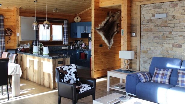 Accogliente interno di una baita con arredamento in legno, zona pranzo, cucina e soggiorno con divano blu e una pelle di animale appesa al muro.