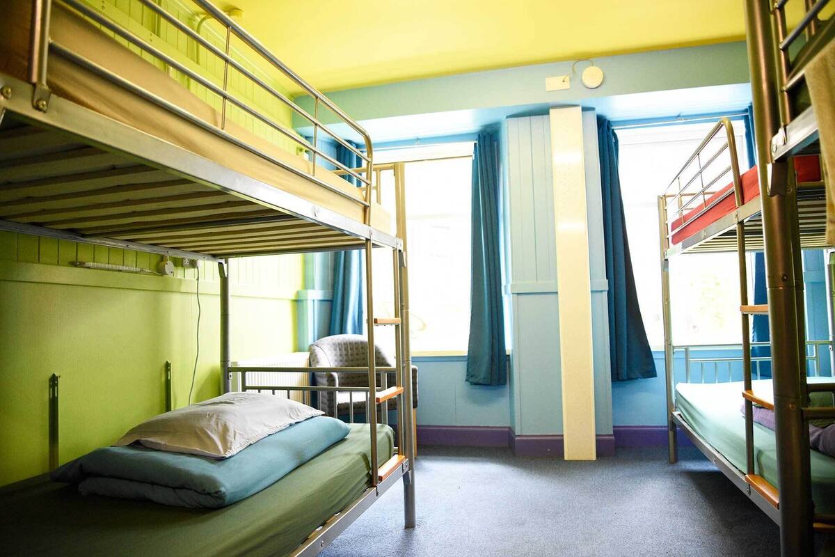 A dormitory room with bunk beds, a small desk, and a window with curtains.