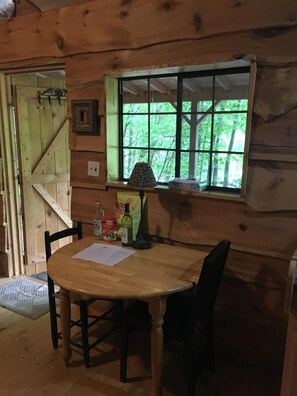 Una cabaña de madera con una mesa de comedor, sillas, una ventana con vistas a follaje y una puerta con diseño triangular.