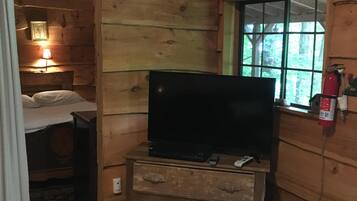 A wooden cabin room with a bed, a television on a dresser, a window, and a heater.
