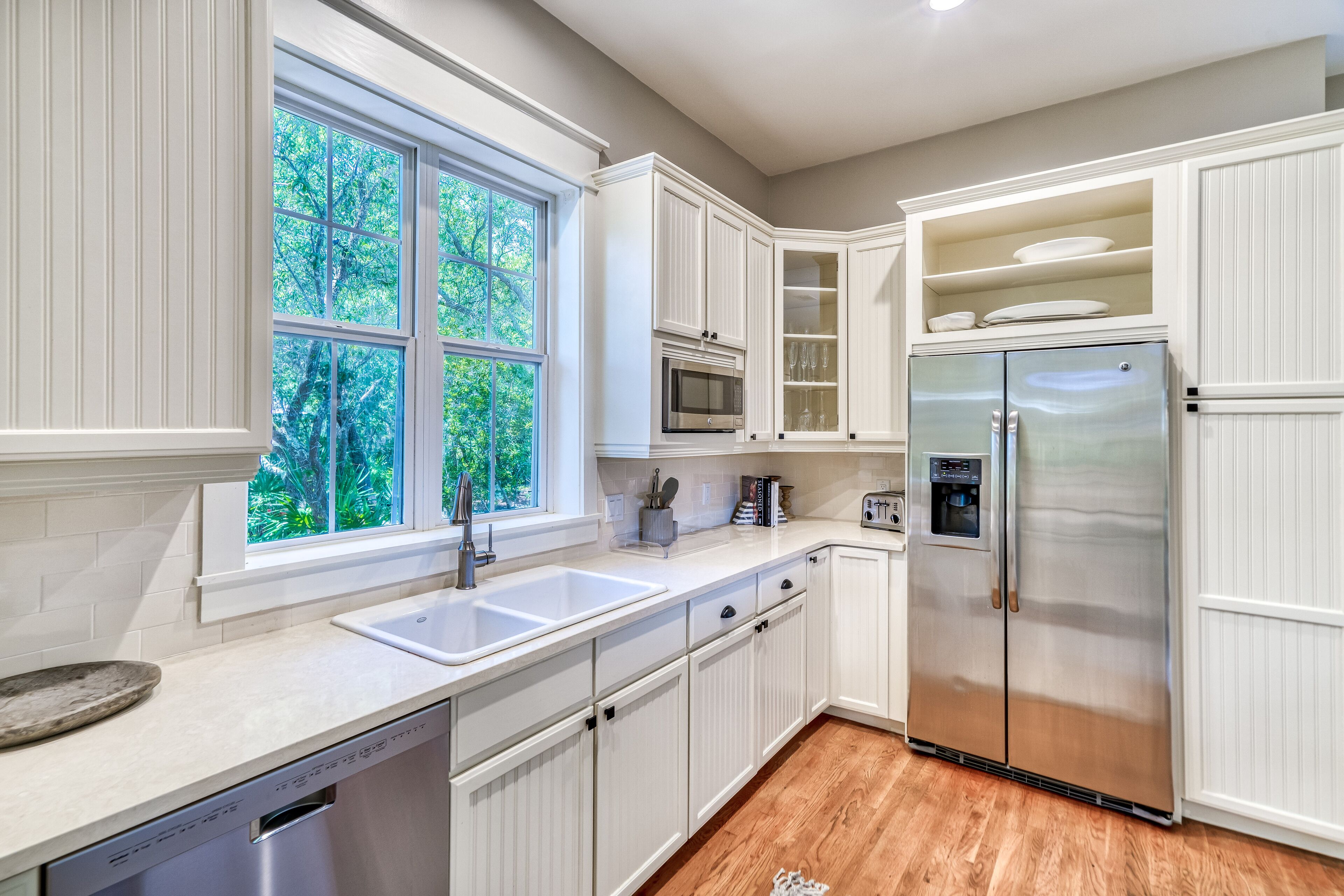 A modern kitchen with white cabinetry, a stainless steel refrigerator, a built-in microwave, and a window with a view of greenery.