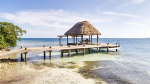Une jetée en bois mène à une cabane à toit de chaume, où se trouvent un banc et une table, le tout surplombant une eau d’un bleu limpide.