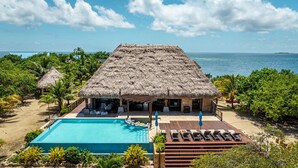 Un bâtiment à toit de chaume doté d’une piscine et d’un espace de détente, entouré de palmiers et offrant une vue sur la mer.