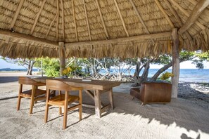 A wooden table and chairs under a thatched roof, with a view of the sea and trees in the background.