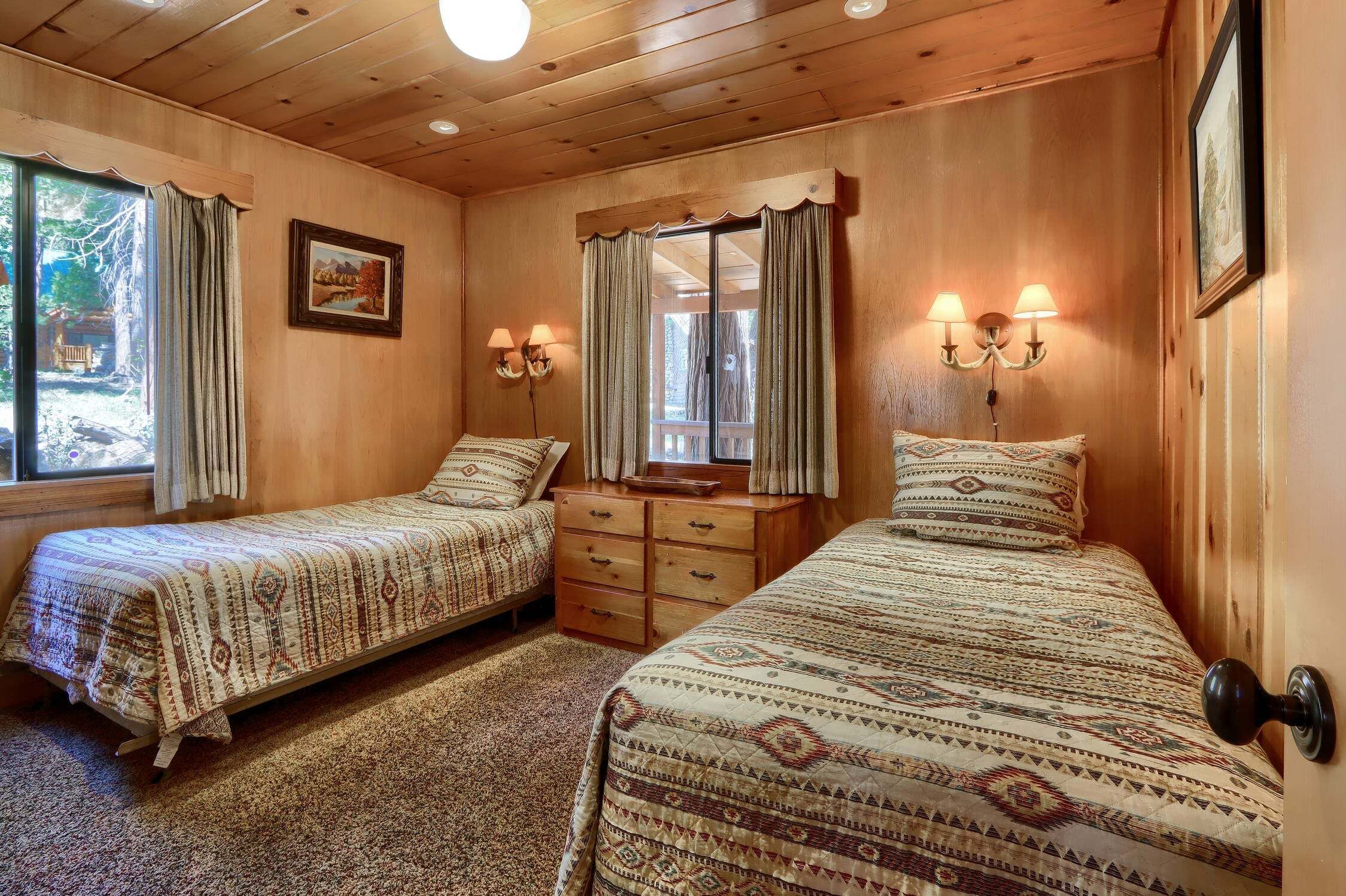 Une chambre avec deux lits, une commode en bois, une fenêtre avec des rideaux et des lampes fixées au mur.