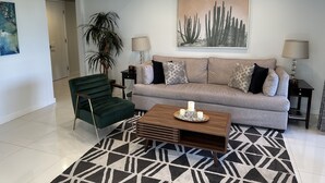 Un salon moderne avec un fauteuil vert, un canapé beige orné de coussins à motifs, une table basse en bois et une grande œuvre d’art représentant un cactus accrochée au mur.