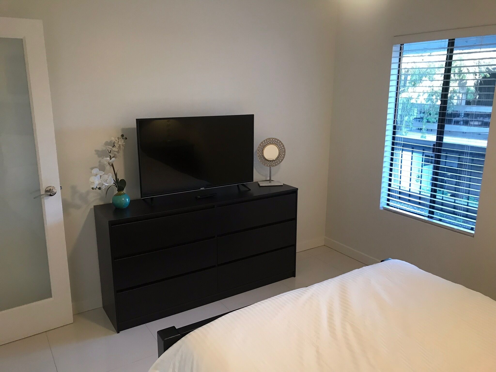 Une chambre à coucher avec un lit, un téléviseur posé sur une commode, une fenêtre avec des stores et une porte.