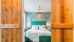 Une chambre à coucher avec une tête de lit recouverte d’un tissu turquoise, un plaid blanc et moelleux, et un cadre de porte en bois.