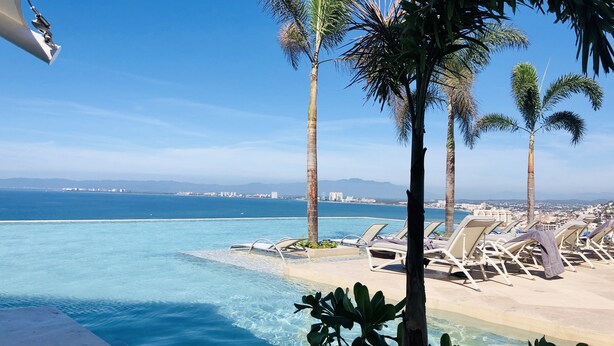Ein Poolbereich mit Liegestühlen und Palmen, mit Blick auf das klare, blaue Meer und eine entfernte Stadtlandschaft.