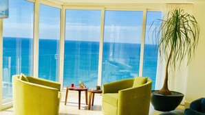 Ein modernes Wohnzimmer mit großen Fenstern mit Meerblick, ausgestattet mit gelben Sesseln, einem kleinen Tisch und einer Topfpflanze.