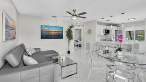 A modern living room with a glass coffee table, a gray sofa, a ceiling fan, and a dining area with a glass table and chairs. - 2BR Hot Tub Oasis - 2 Mi FLL, 4 Mi Beach, Sleeps 6 (Dania Beach)