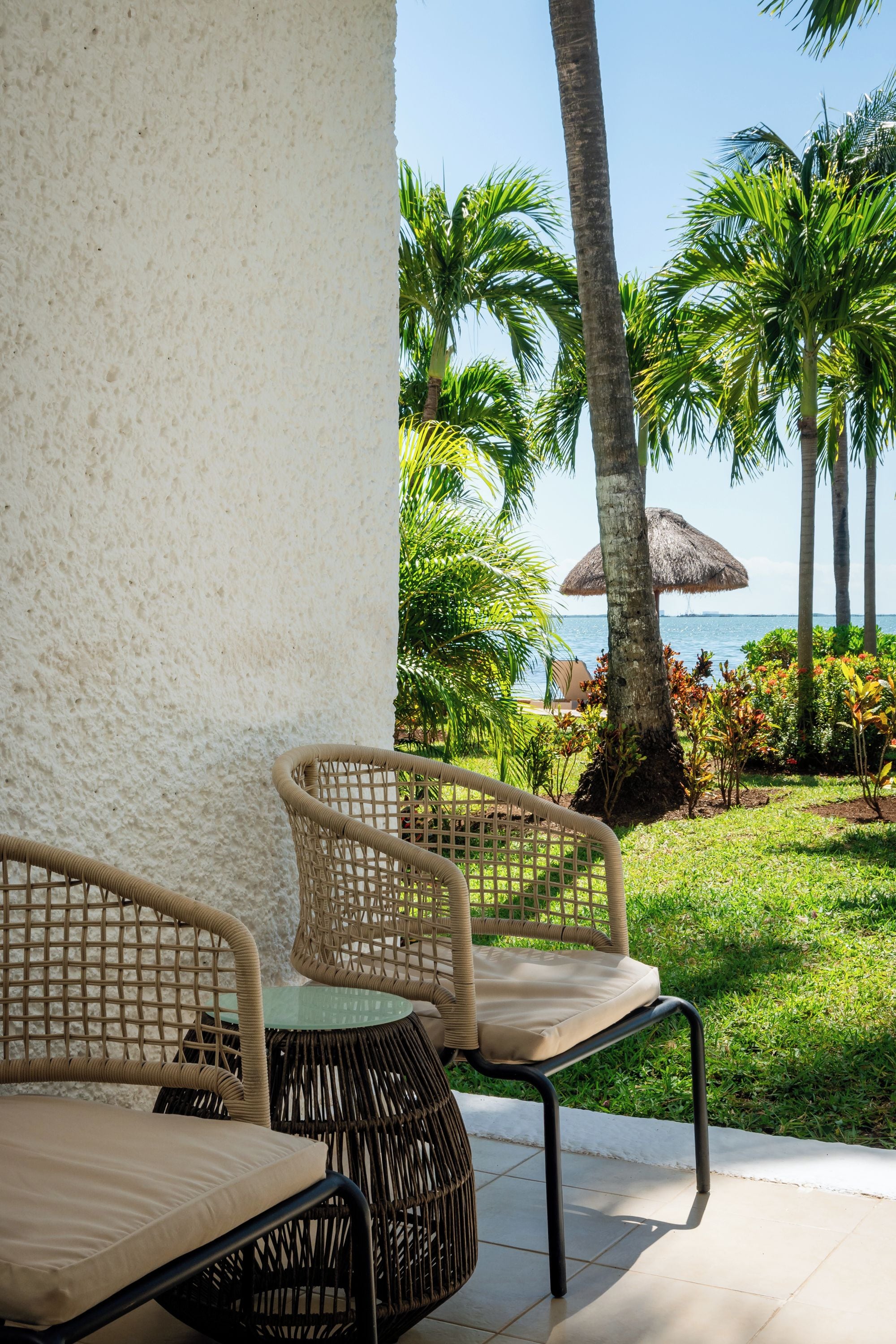 Wicker chairs with cushions, a thatched umbrella, and a view of the sea with palm trees.