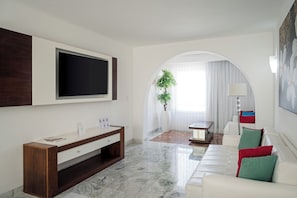 Un salon moderne doté d’un téléviseur à écran plat, d’une console en bois, d’un canapé blanc agrémenté de coussins colorés et d’une grande fenêtre avec des rideaux.