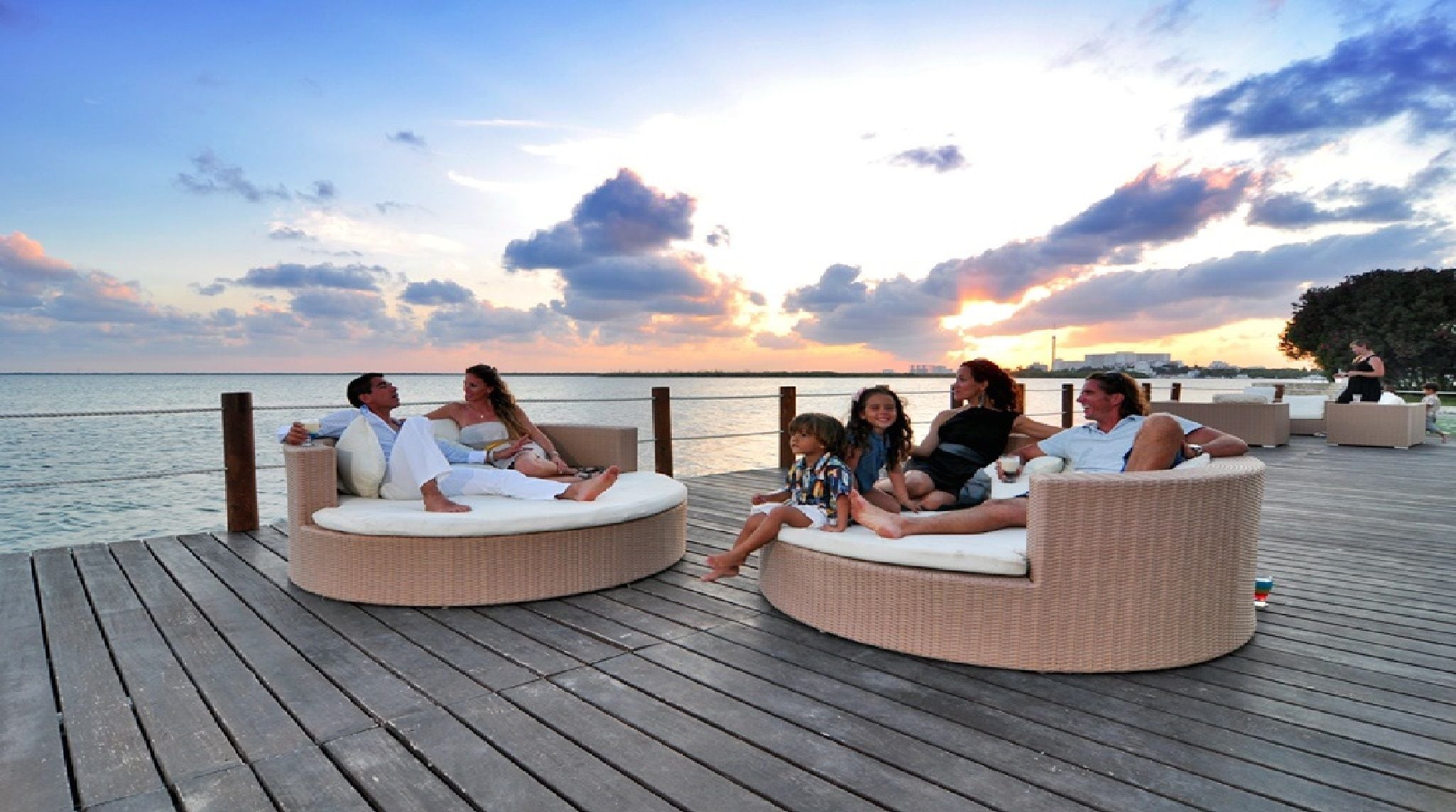 A group of people relaxing on wicker lounge chairs on a wooden deck overlooking a body of water during sunset.