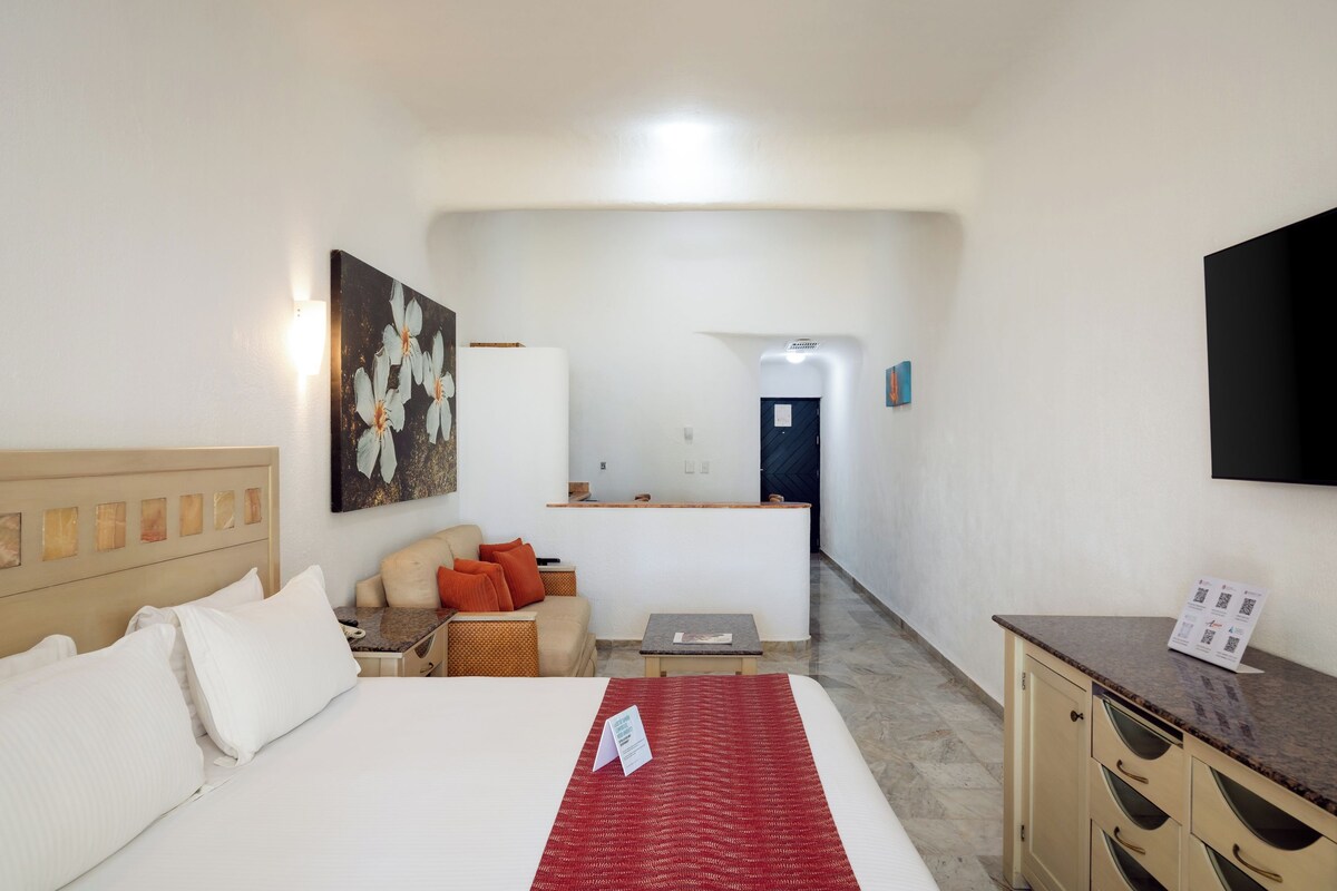 A hotel room with a bed, a desk, a chair, a small table, a sofa, and a painting on the wall.