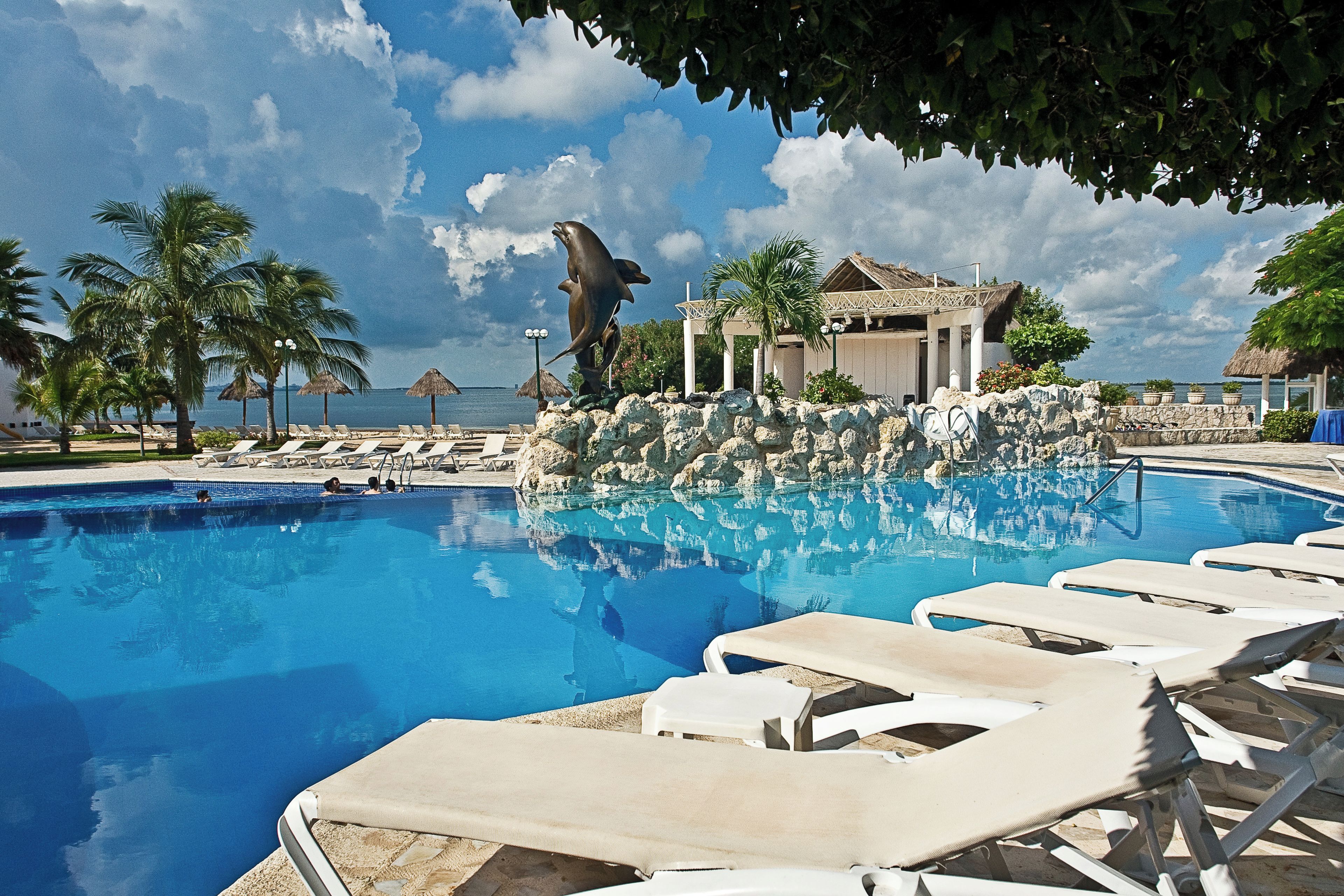 A swimming pool with lounge chairs, a dolphin statue, and a building with a thatched roof.