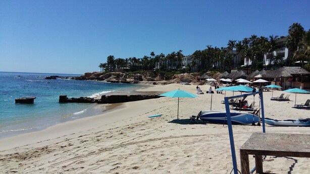 Una playa con agua azul cristalina, orilla de arena y palmeras. Hay sombrillas y sillas de descanso para tomar el sol.