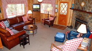 Un salon chaleureux avec un canapé en cuir marron, un fauteuil bleu et une table basse en bois. On y trouve un téléviseur posé sur un meuble en bois, une cheminée et un mur en pierre.