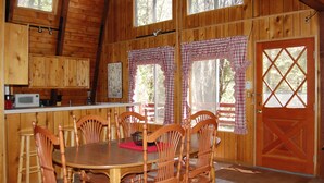 L’intérieur d’un chalet en bois comprend une table à manger, des chaises, un coin cuisine et de grandes fenêtres ornées de rideaux à carreaux.