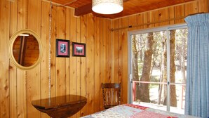 Une pièce de style cabane en bois, comprenant un lit, une table, une chaise, une fenêtre donnant sur la forêt et deux tableaux encadrés accrochés au mur.