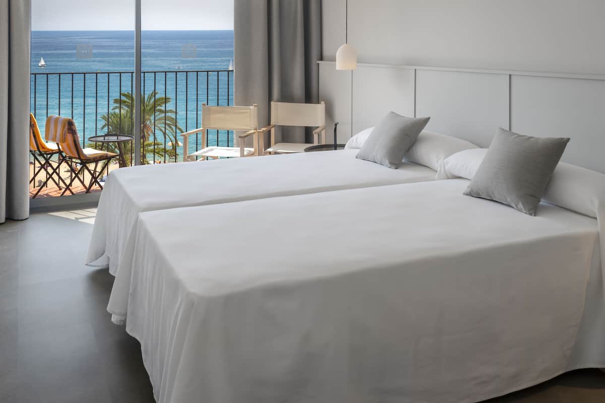 Een hotelkamer met twee bedden, een balkon met uitzicht op zee en een zithoek met stoelen en een tafel.