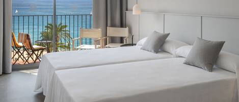 A hotel room with two beds, a balcony with a view of the sea, and a seating area with chairs and a table.