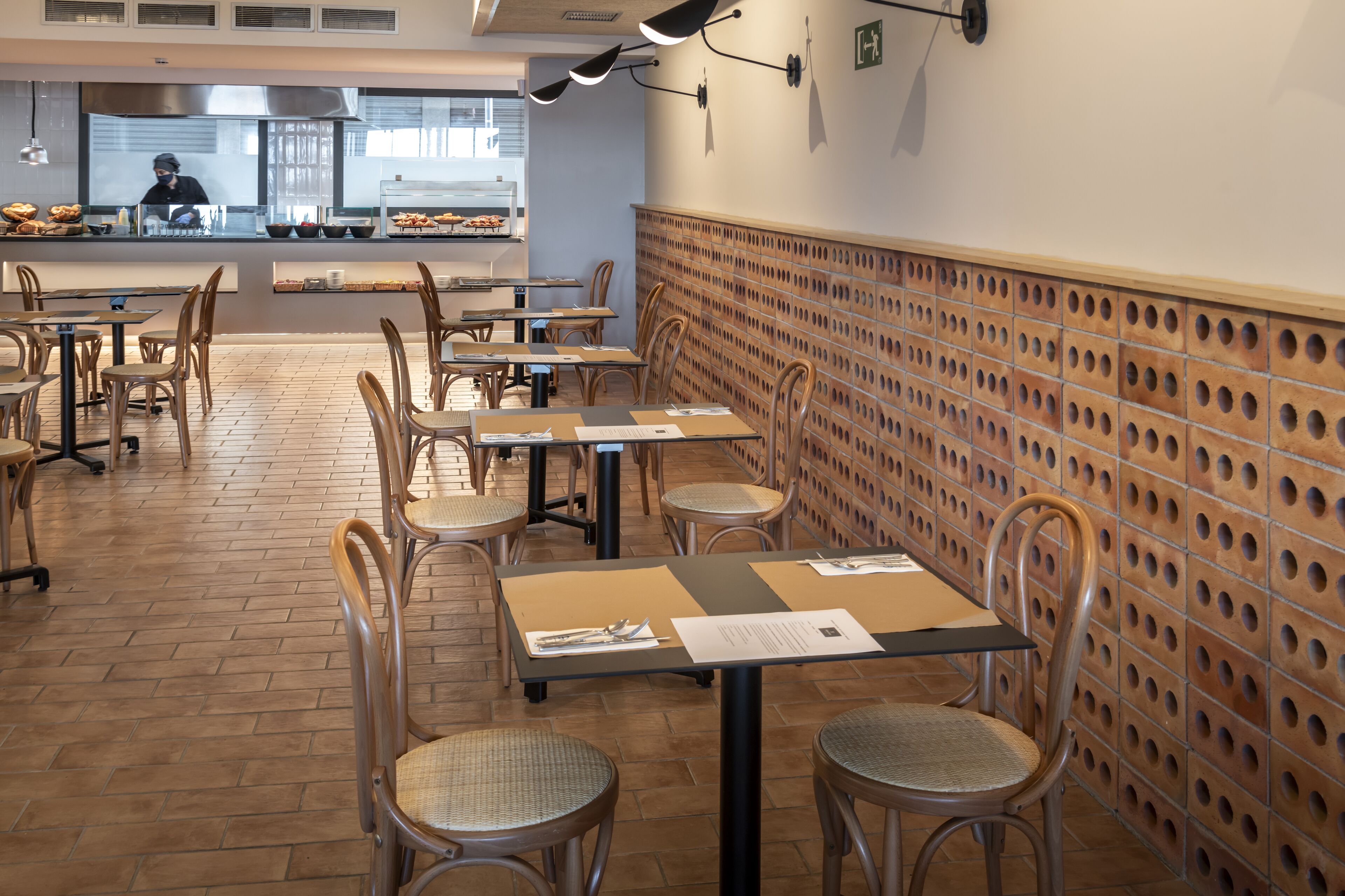 A restaurant interior with wooden tables and chairs, a kitchen in the background, and a patterned wall.