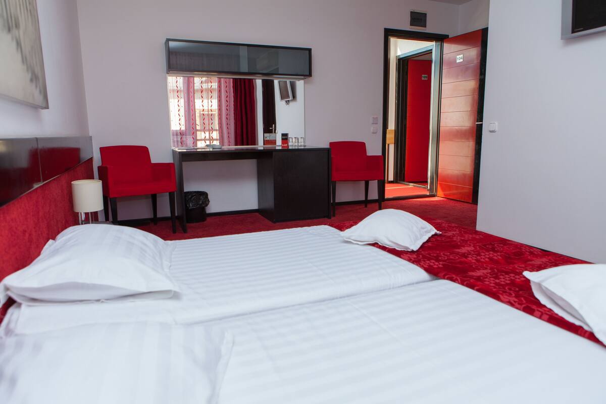 Habitació d'hotel amb un llit, escriptori, dues cadires vermelles, televisió i un passadís amb moqueta vermella.