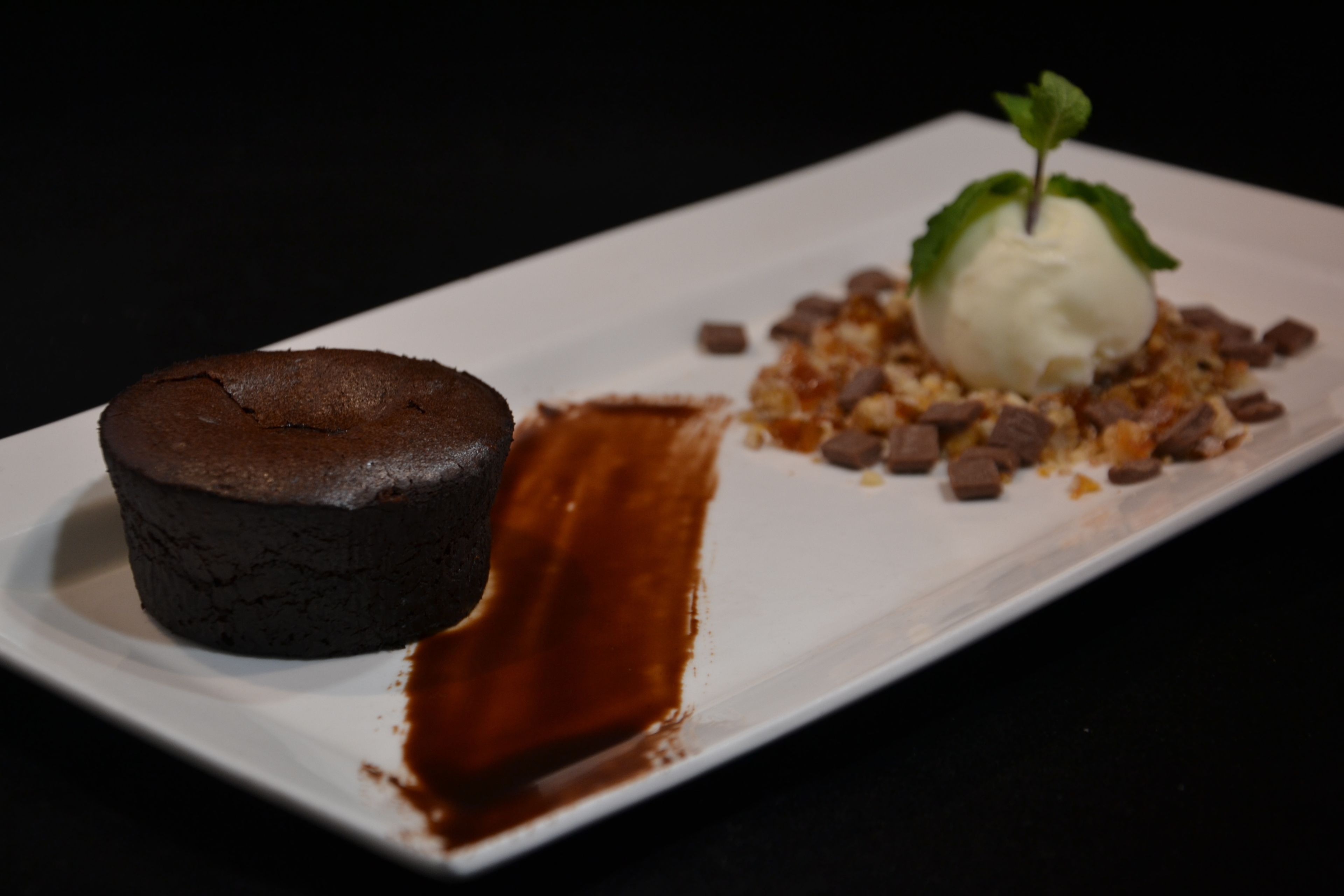 A dessert plate with a chocolate cake and a scoop of ice cream, garnished with chocolate pieces and a mint leaf.