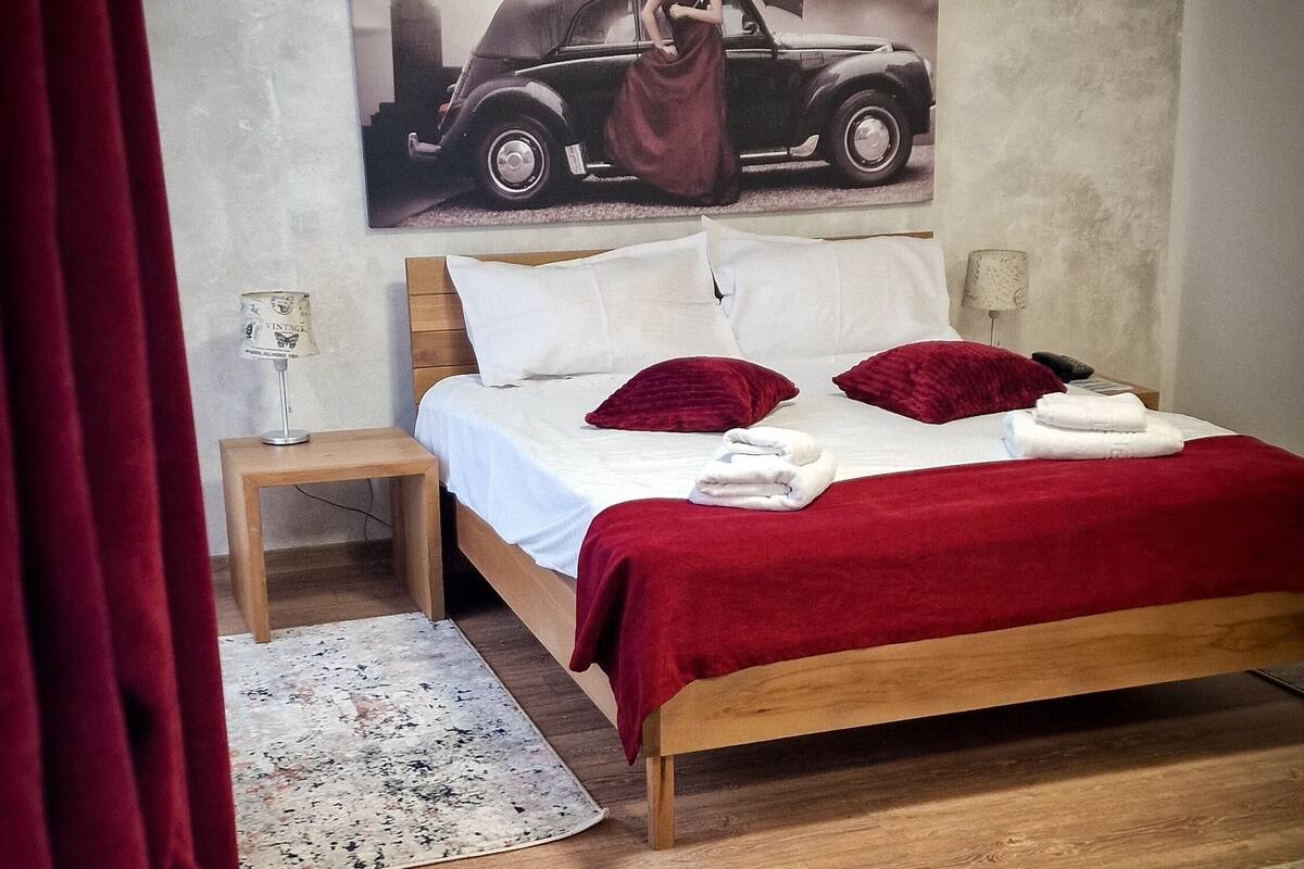 Un dormitori amb un llit, una taula de nit, una làmpada, una cortina vermella i un pòster d'un cotxe a la paret.