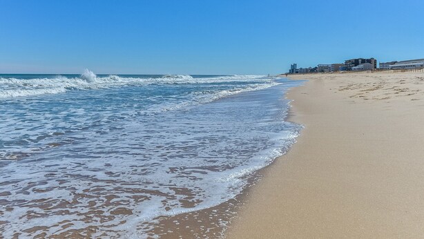 Una playa con olas, una orilla arenosa y edificios al fondo.