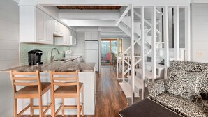 Cocina moderna con armarios blancos, encimera de mármol y cafetera. Zona de comedor de madera con dos sillas y un sofá estampado. Una escalera blanca conduce a otra habitación.