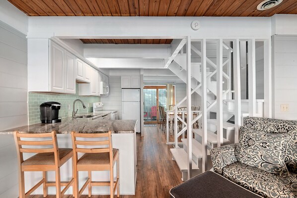 A modern kitchen with white cabinets, a marble countertop, and a coffee maker. There is a wooden dining area with two chairs and a patterned sofa. A white staircase leads to another room.