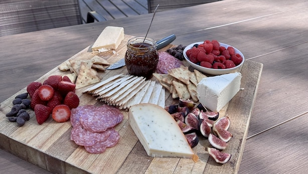 Tábua de madeira com diversos queijos, frutas e bolachas sobre uma mesa ao ar livre.