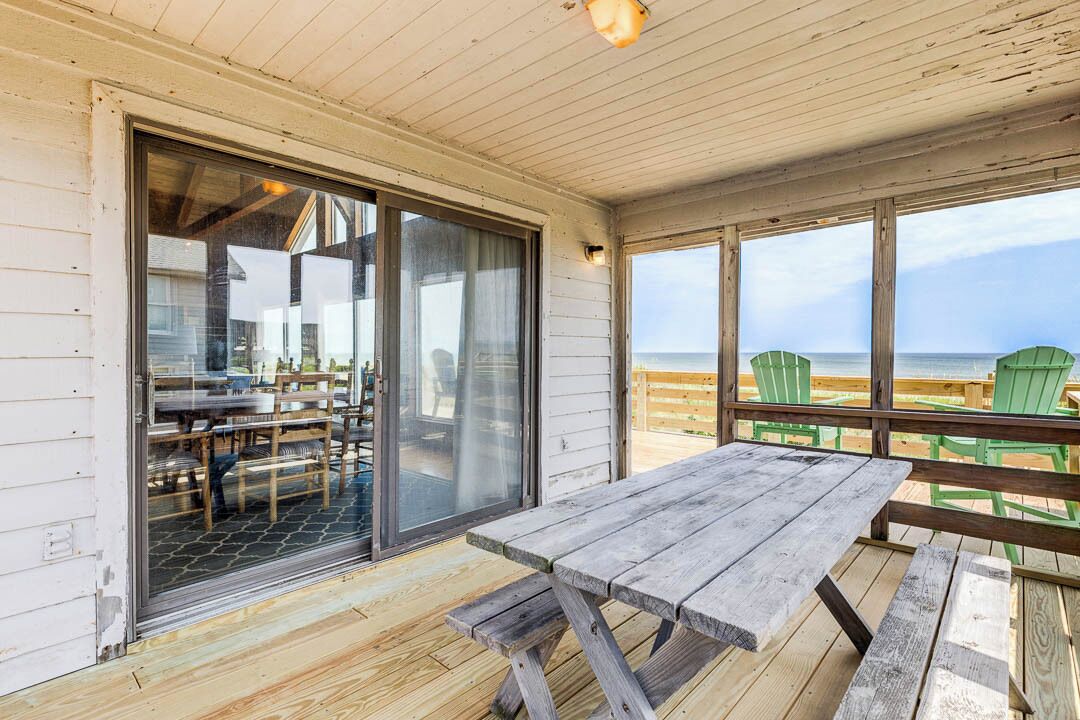 A covered outdoor area with a wooden table and chairs, a sliding glass door leading to a patio, and a view of the beach.