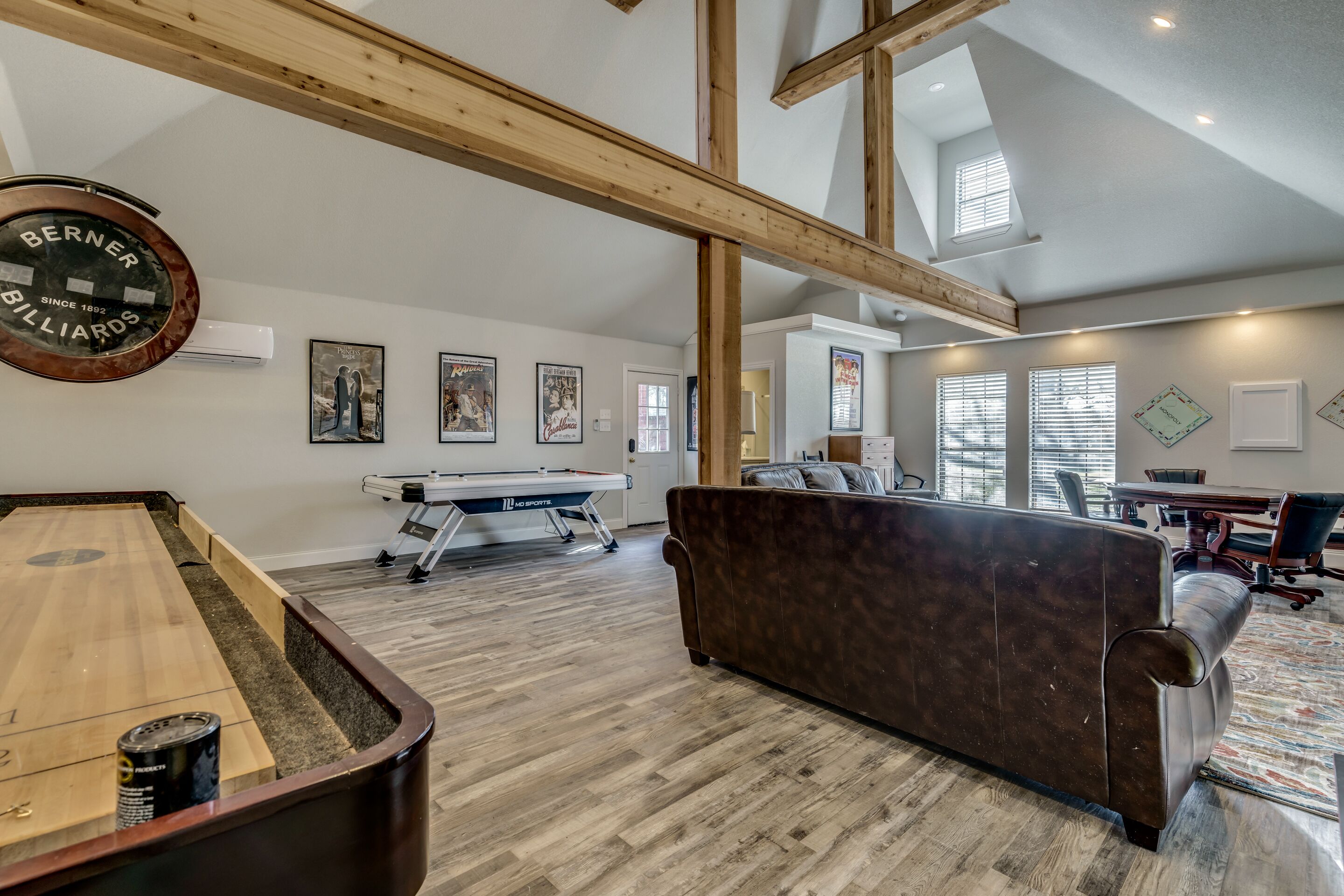 A billiards table, a pool table, and a brown sofa in a room with wooden beams and large windows.