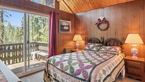 A bedroom with a large bed, wooden walls, a wooden ceiling, a view of trees, a bench, and a table outside the window.